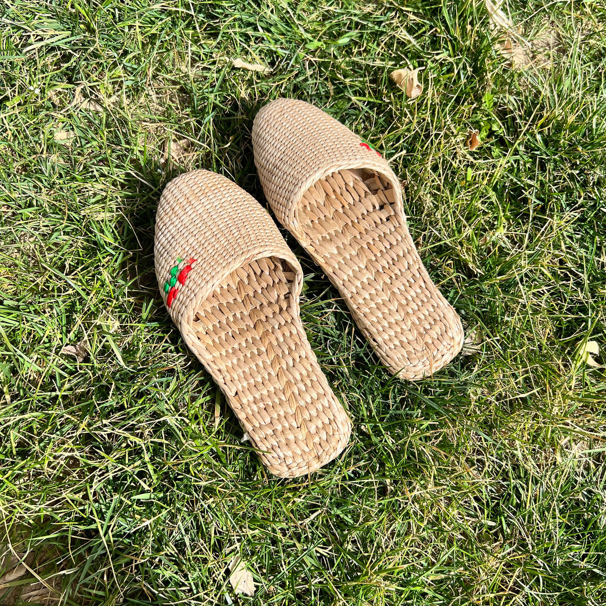 Pair of handmade straw slippers with closed toe and embroidered red and green cherry detail, placed on green grass. Natural beige woven texture, lightweight and breathable, ideal for summer wear.