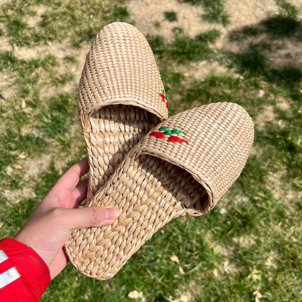 Hand holding a pair of handmade straw slippers with closed toe and embroidered red and green cherry detail. Natural beige woven texture, lightweight and breathable, ideal for house shoes or summer sandals.