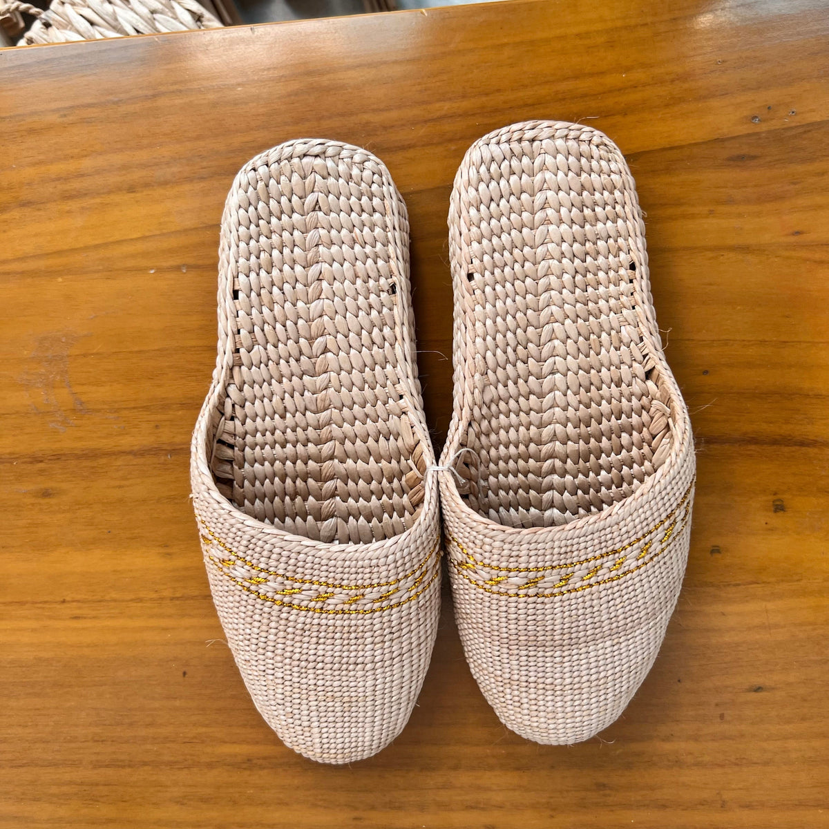 Top view of beige woven straw slippers with yellow decorative trim, placed on a wooden surface. Features breathable natural fiber texture, lightweight soft sole, ideal for summer beach wear or casual indoor use.