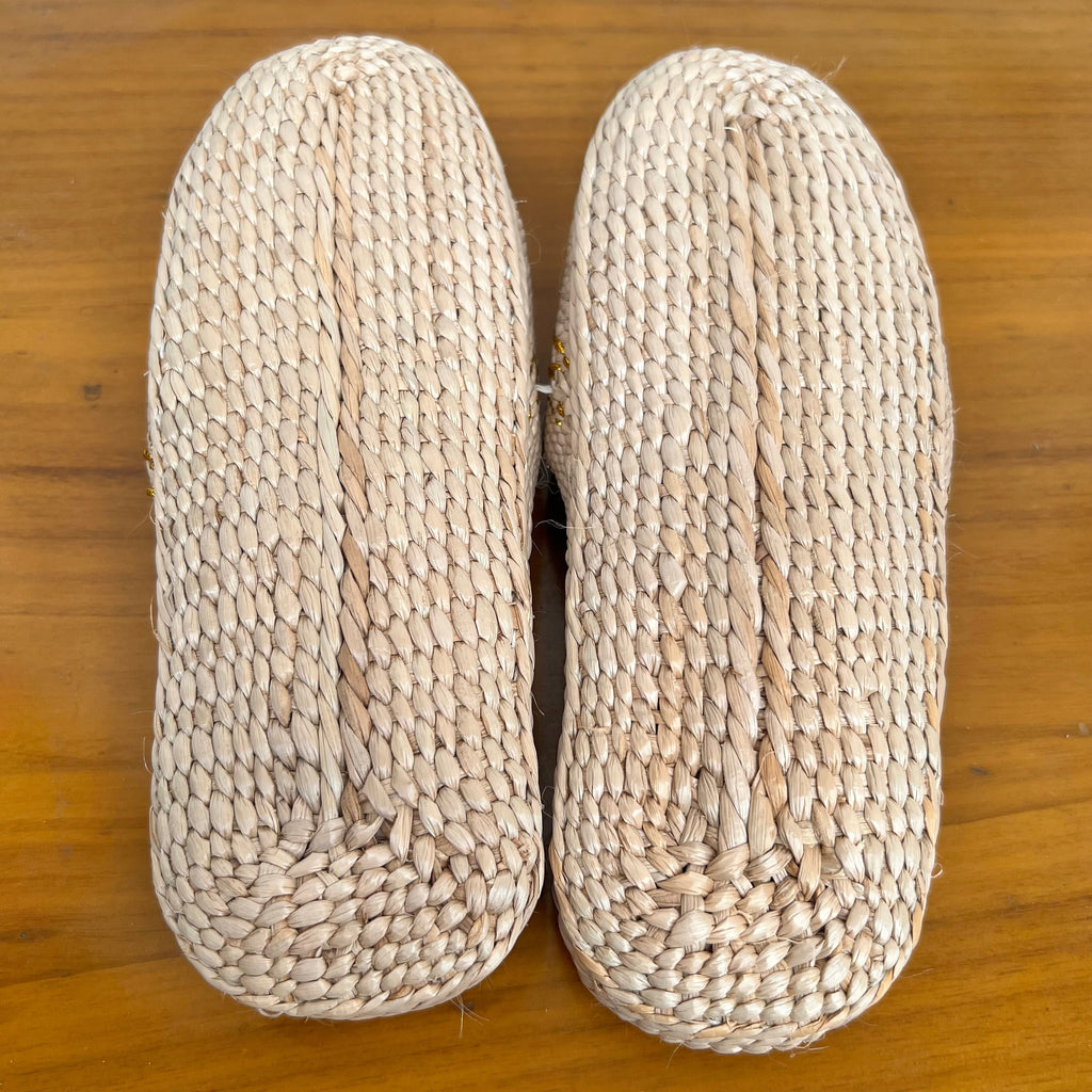 Bottom view of beige woven straw slippers showing tightly braided natural fibers. Features flat, lightweight sole designed for breathable comfort, ideal for summer beach wear or indoor casual use.