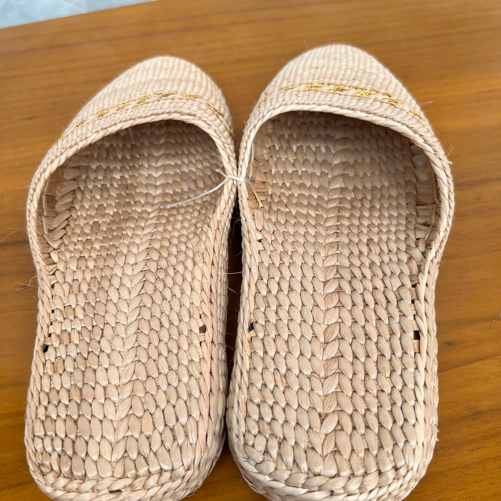 Close-up top view of beige woven straw slippers with soft flat soles and subtle yellow decorative trim. Made from breathable natural fibers, lightweight and comfortable for summer beach wear or indoor casual use.