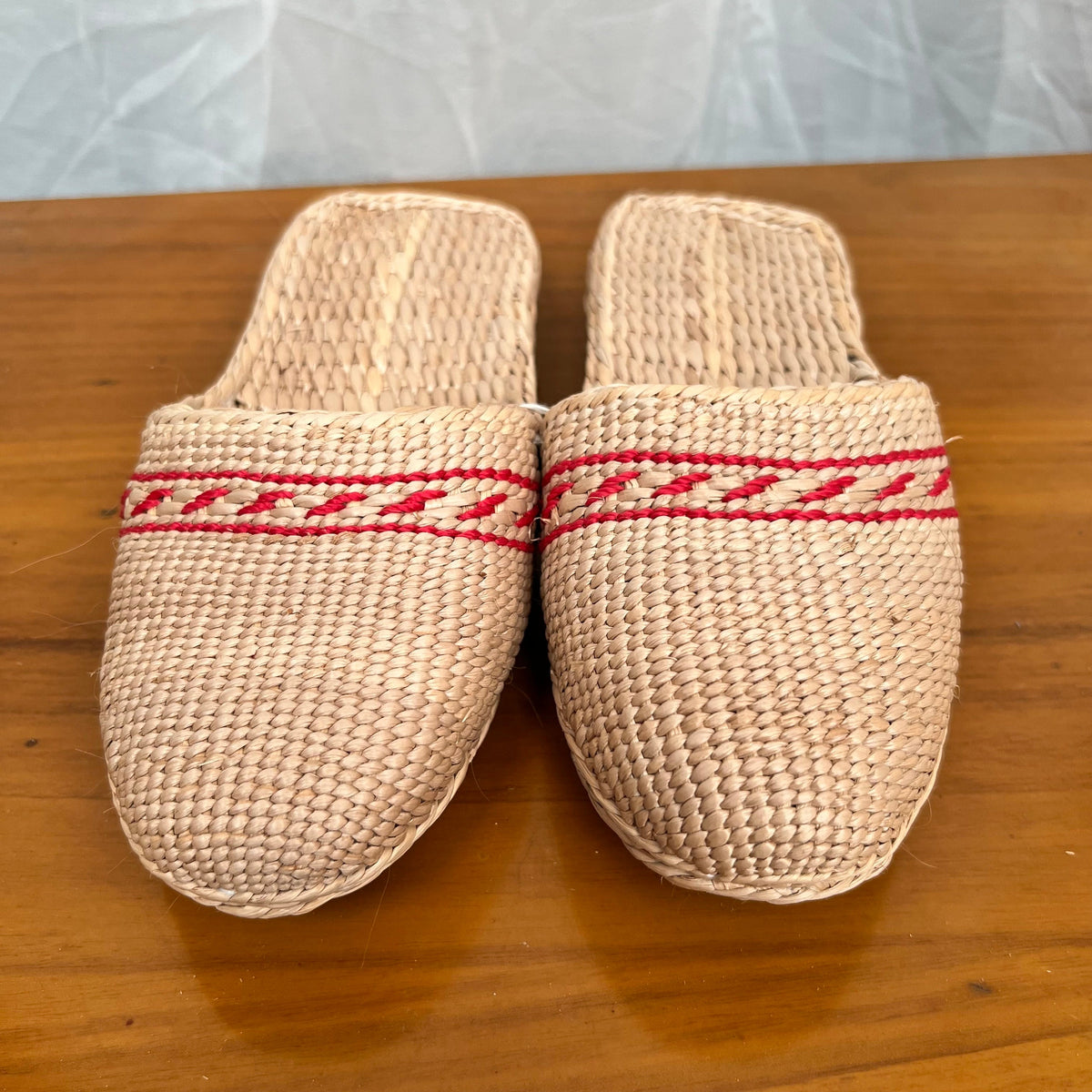 Front view of beige woven straw slippers with red decorative trim across the uppers, displayed on a wooden surface. Features flat braided soles, lightweight breathable texture, ideal for summer beach wear or indoor casual comfort.