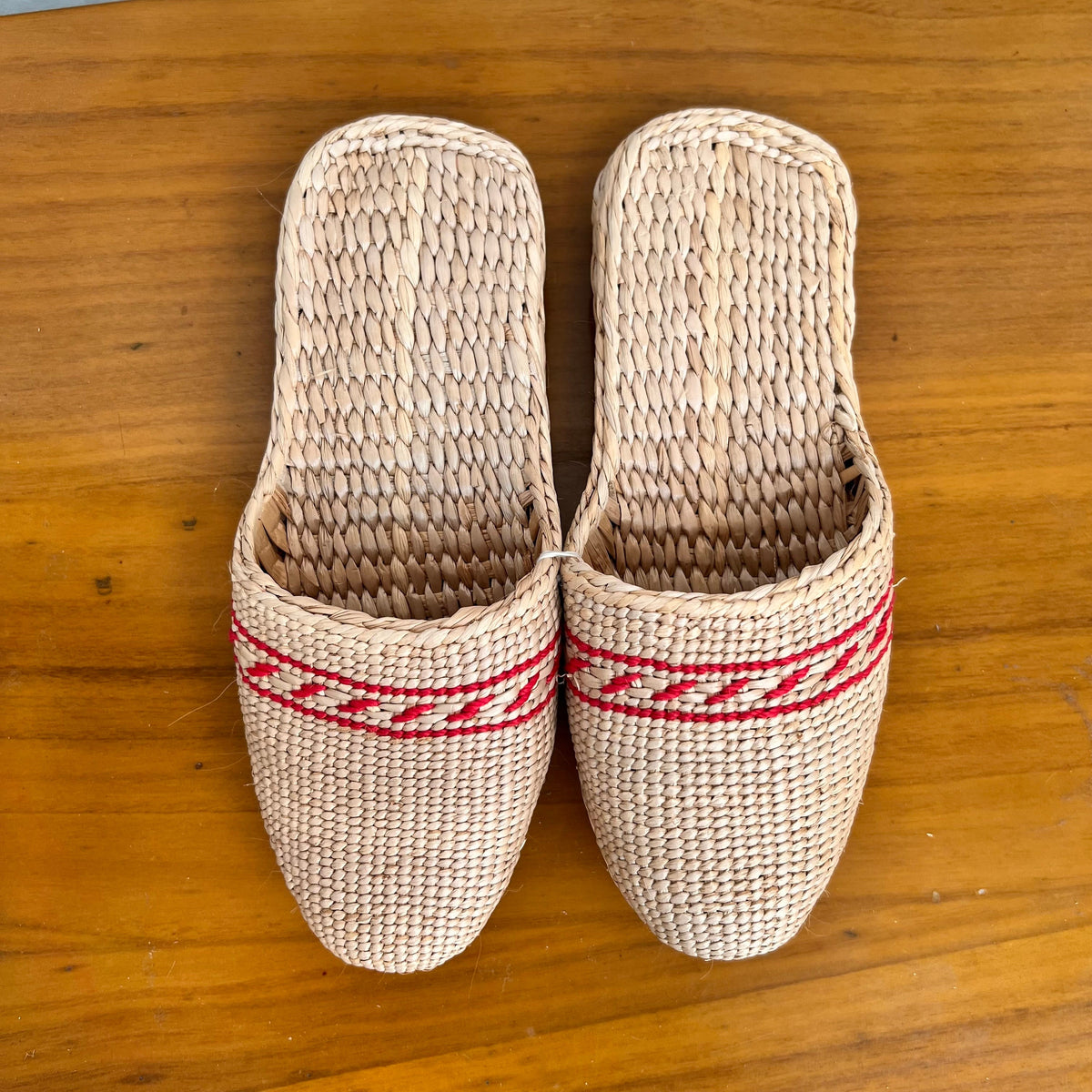 Top view of woven straw slippers in beige with red decorative trim, placed on a wooden surface. Features flat braided soles, breathable natural fibers, lightweight and comfortable for summer beach or indoor wear.
