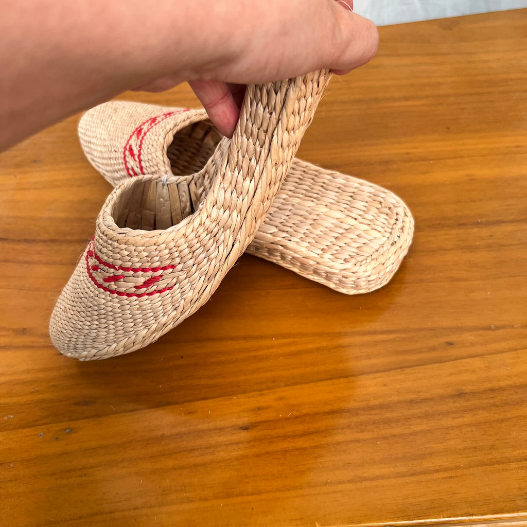 Hand holding a woven straw slipper in beige with red decorative trim, showing flexible flat braided sole. Second slipper rests on wooden surface. Lightweight, breathable, eco-friendly design ideal for summer beach or indoor wear.
