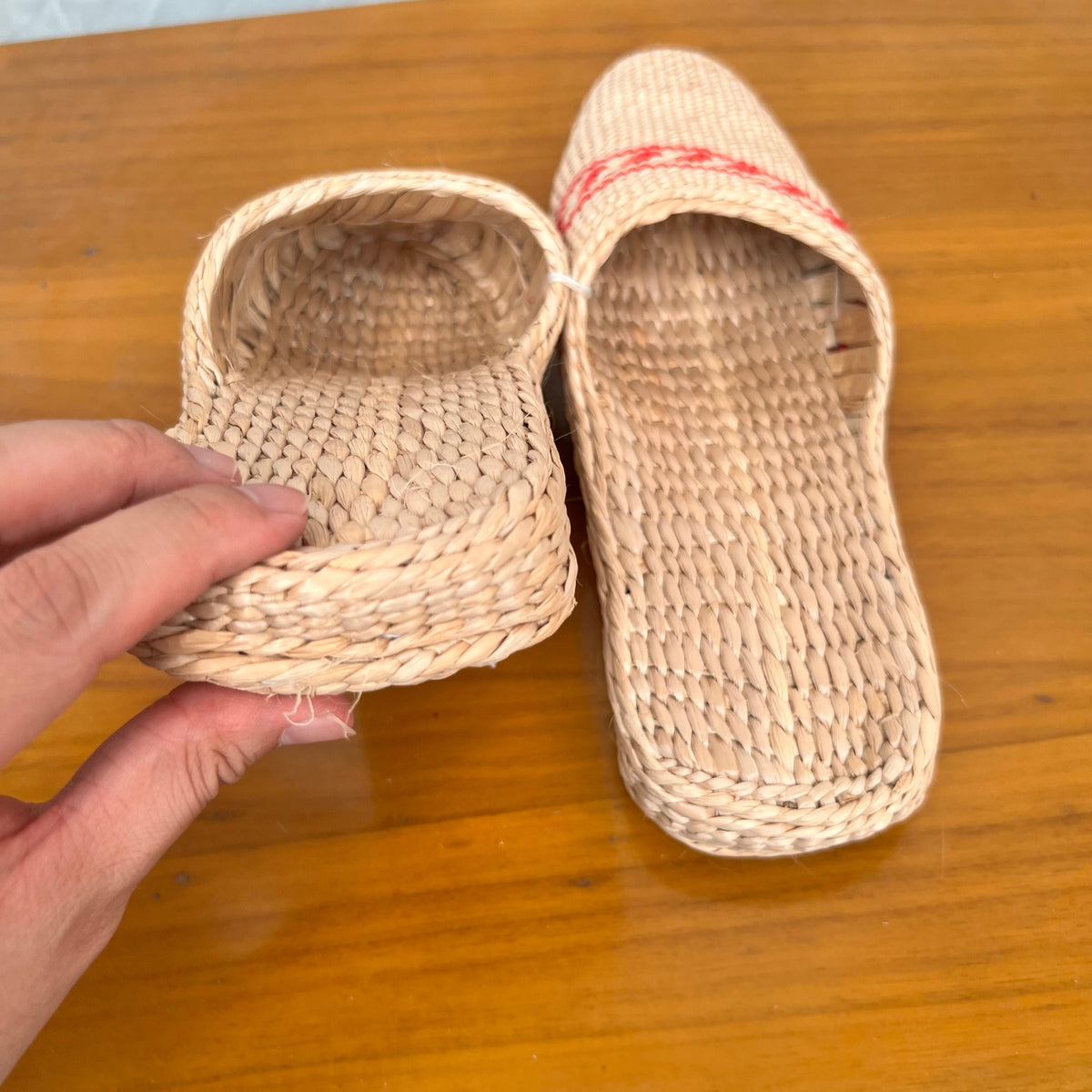 Close-up of woven straw slippers with flat braided soles, one being held to show thickness. Beige natural fiber design with red decorative trim, lightweight and breathable, ideal for summer beach wear or indoor casual comfort.