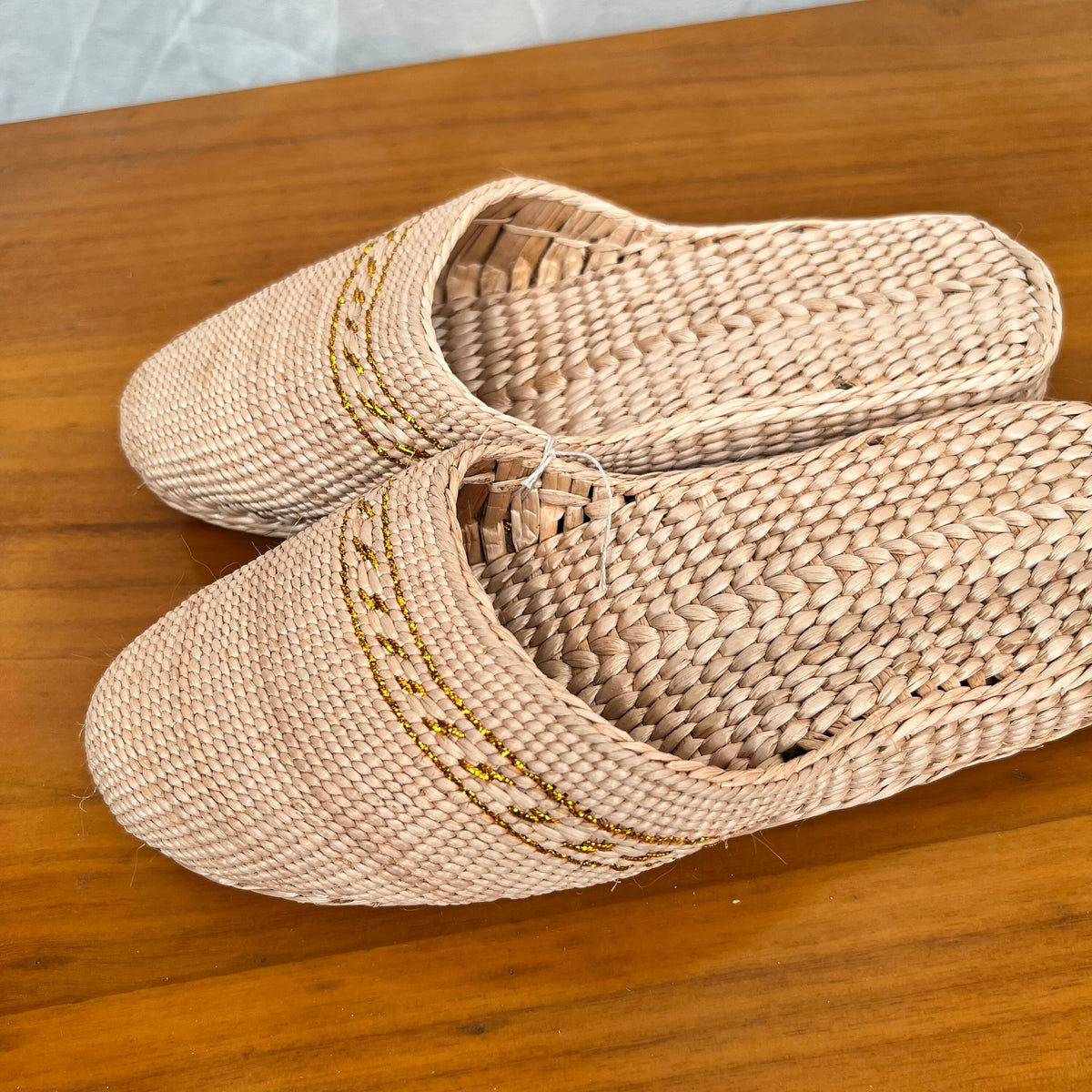 Pair of beige woven straw slippers with yellow decorative trim, displayed on a wooden surface. Features soft sole, lightweight design, and breathable texture for comfortable summer beach or indoor wear.