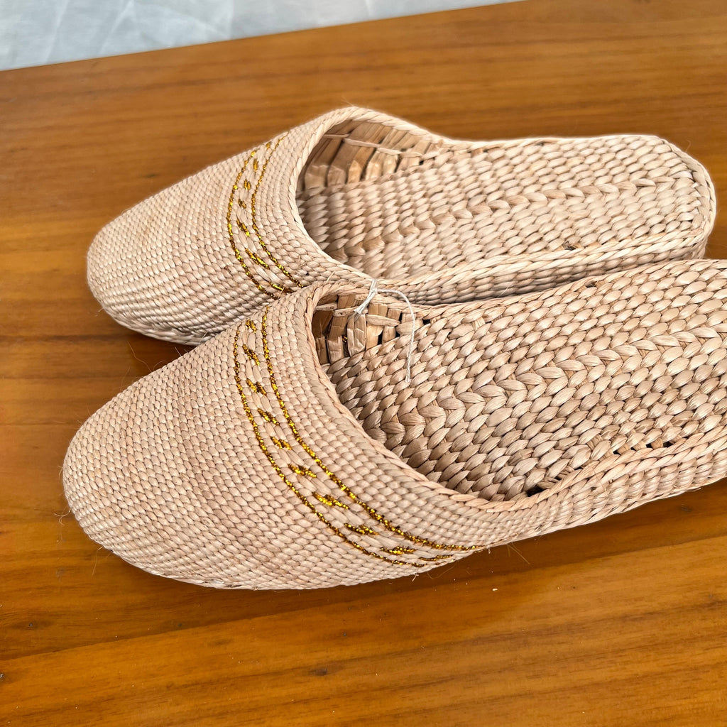 Pair of beige woven straw slippers with yellow decorative trim, displayed diagonally on a wooden surface. Features breathable natural fiber texture, soft lightweight sole, ideal for summer beach wear or indoor casual use.