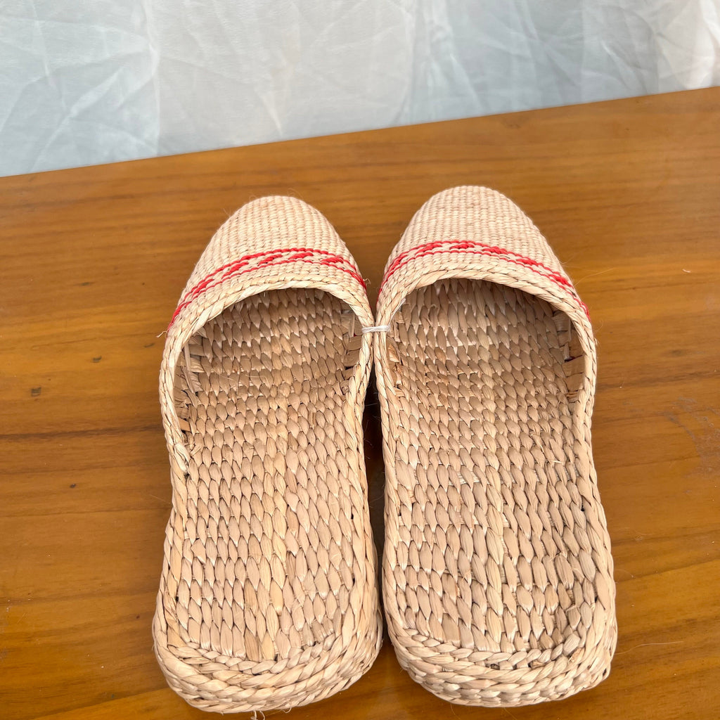 Front view of woven straw slippers in beige with red decorative trim, placed on a wooden surface. Features flat braided soles, breathable natural fiber design, lightweight and comfortable for summer beach or indoor wear.