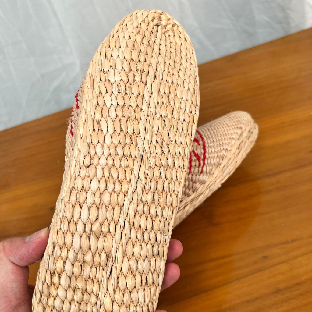 Close-up of the bottom sole of woven straw slippers in beige, showing tightly braided natural fibers. A hand holds one slipper to display the sole detail. Lightweight, breathable, eco-friendly design with red decorative trim on the uppers.
