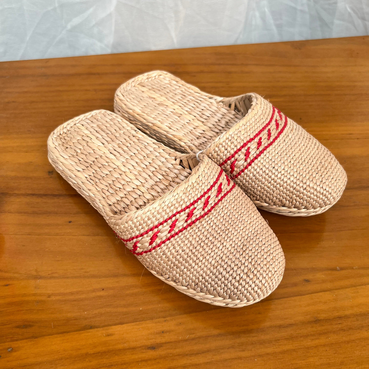 Pair of woven straw slippers in beige with red decorative trim, displayed on a wooden surface. Features flat braided soles, lightweight and breathable design, perfect for summer beach wear or casual indoor comfort.
