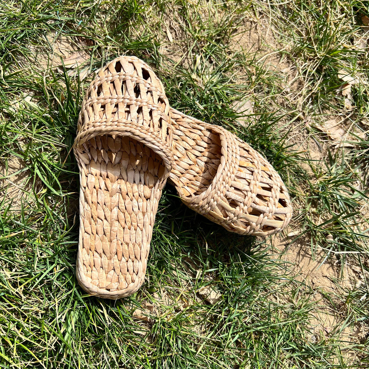 Pair of handmade woven straw sandals with open-weave uppers and flat natural soles, placed on outdoor grass. Lightweight, breathable, eco-friendly design perfect for summer beach wear or casual outdoor activities.
