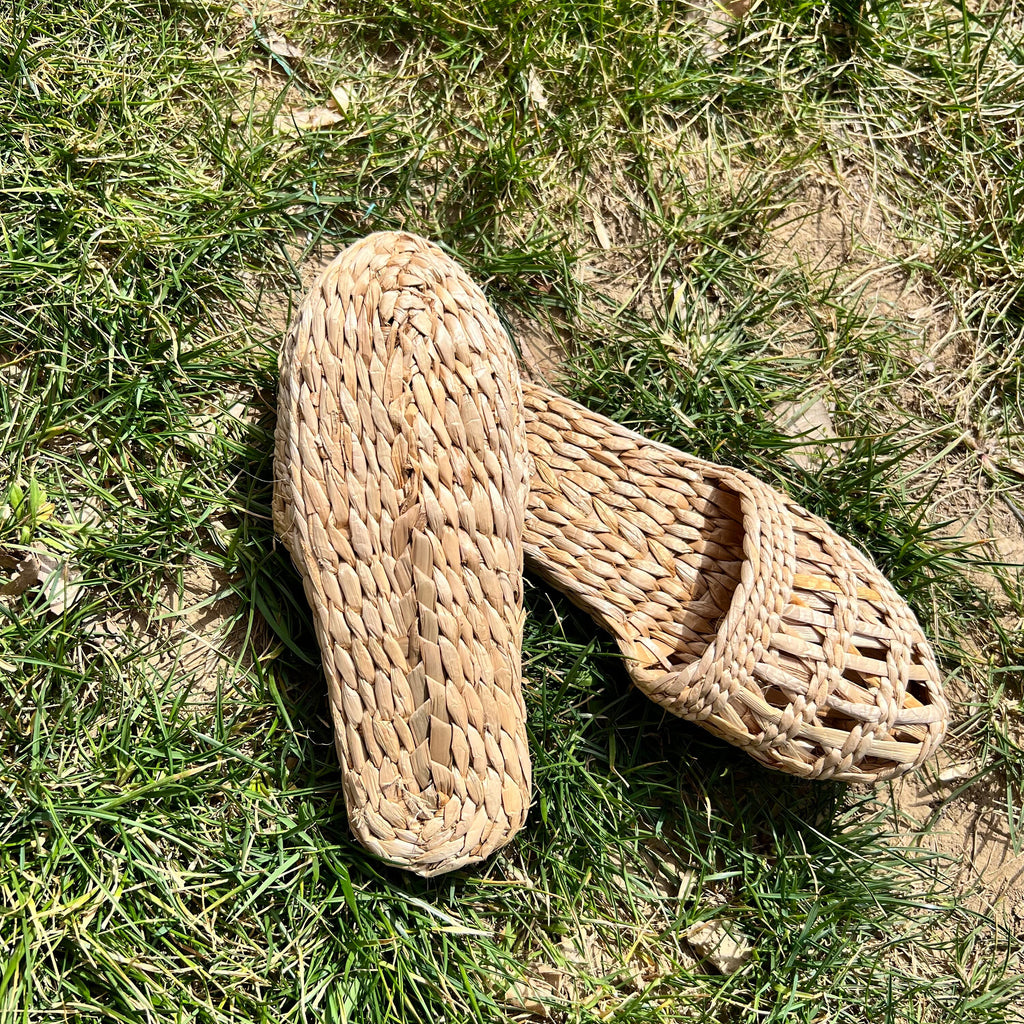 Pair of woven straw slippers with open-weave uppers and flat braided soles, one turned to show the bottom, placed on outdoor grass. Lightweight, breathable, eco-friendly design ideal for summer beach wear or casual outdoor comfort.