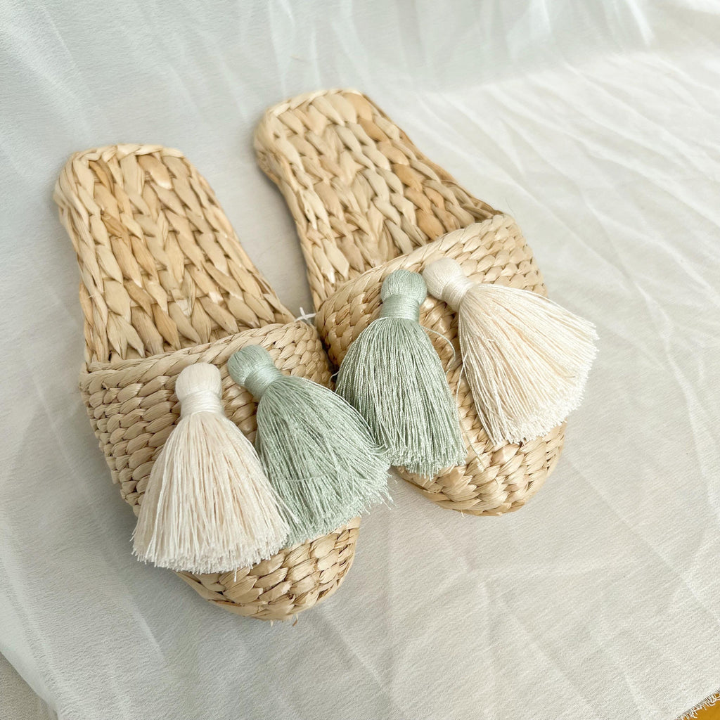 Pair of handmade straw slippers with closed toe and wide woven upper, decorated with three large tassels in white, mint green, and cream. Natural beige color, lightweight and breathable.