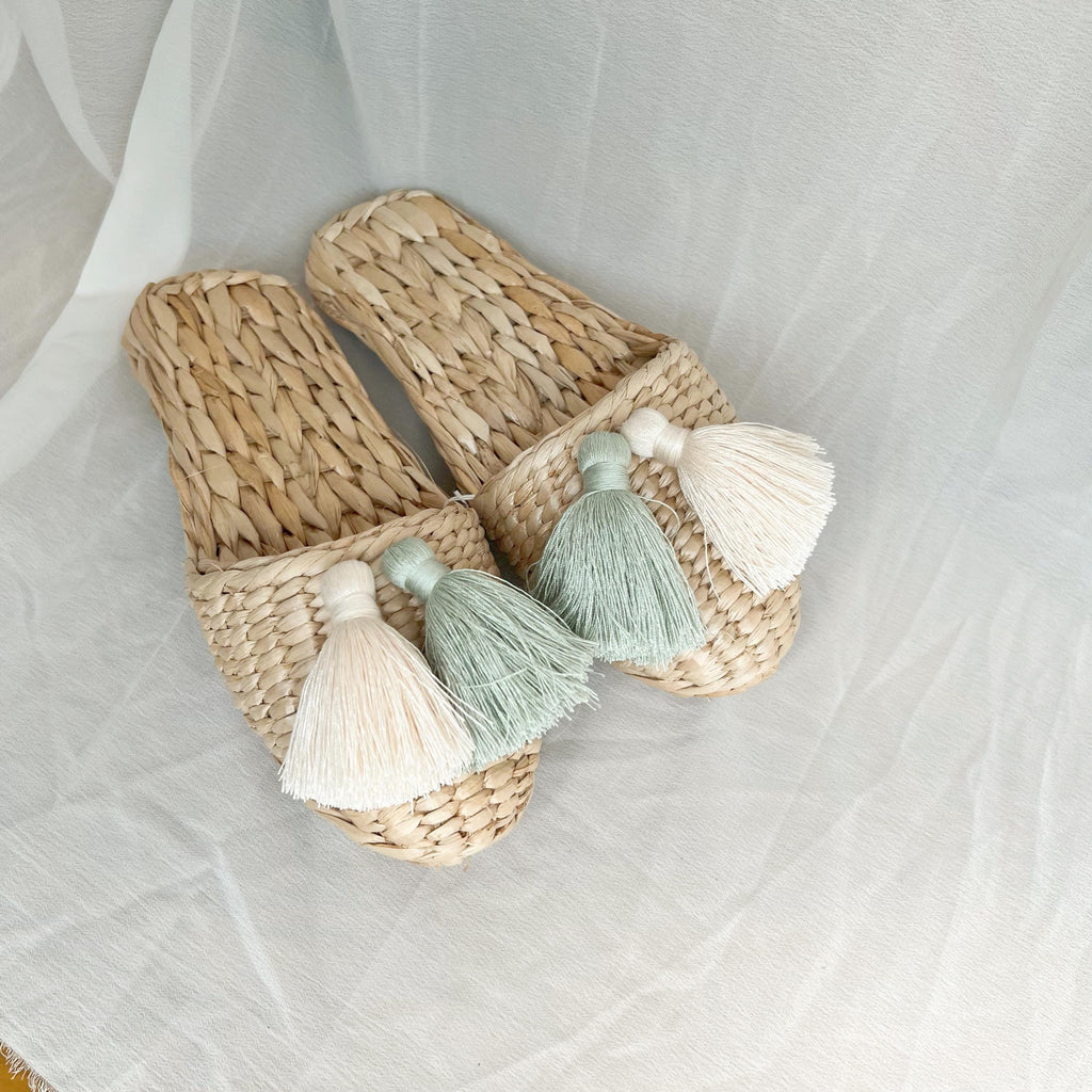 Pair of handmade straw slippers with closed toe and wide woven upper, decorated with three large tassels in cream, mint green, and white. Natural beige color, lightweight and breathable.