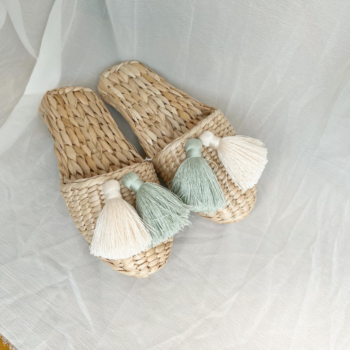 Pair of handmade straw slippers with closed toe and wide woven upper, decorated with three large tassels in cream, mint green, and white. Natural beige color, lightweight and breathable.