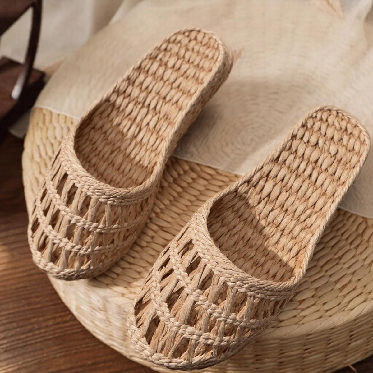 Pair of handmade woven straw slippers with open-weave uppers and tightly braided soles, displayed on a round woven mat. Lightweight, breathable, eco-friendly design ideal for summer beach wear or casual indoor comfort.