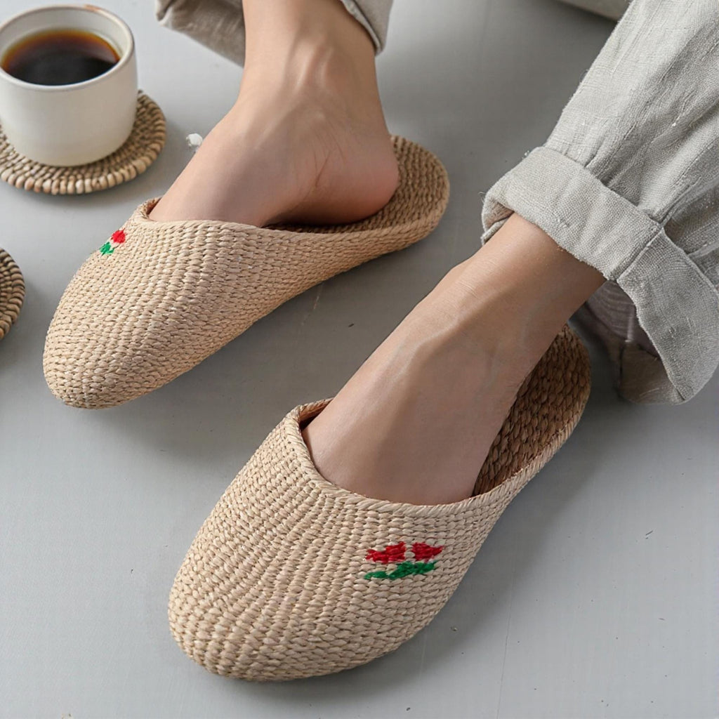 Person wearing handmade straw slippers with closed toe and embroidered red and green cherry detail, paired with rolled-up light pants. Natural beige woven texture, lightweight and breathable.