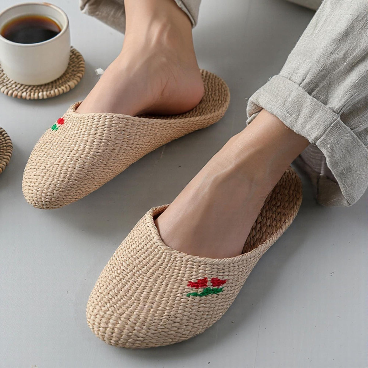 Person wearing handmade straw slippers with closed toe and embroidered red and green cherry detail, paired with rolled-up light pants. Natural beige woven texture, lightweight and breathable.