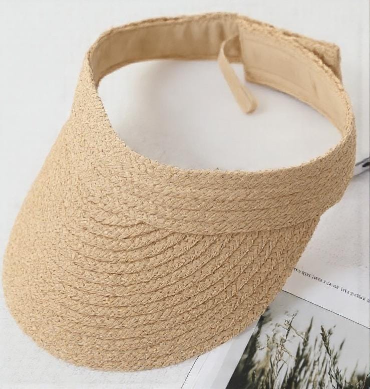 Straw sun hat visor with a wide brim and natural woven texture, resting on a magazine. Lightweight, stylish summer cap for beach, outdoor UV protection, vacation, honeymoon, garden, or as a thoughtful gift for her mom.