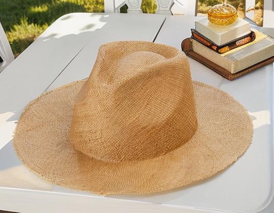 Wide brim straw sun hat with natural woven texture placed on a white outdoor table beside stacked books and sunlight. Lightweight, stylish summer hat for beach, outdoor UV protection, vacation, honeymoon, garden, or as a thoughtful gift for her mom.