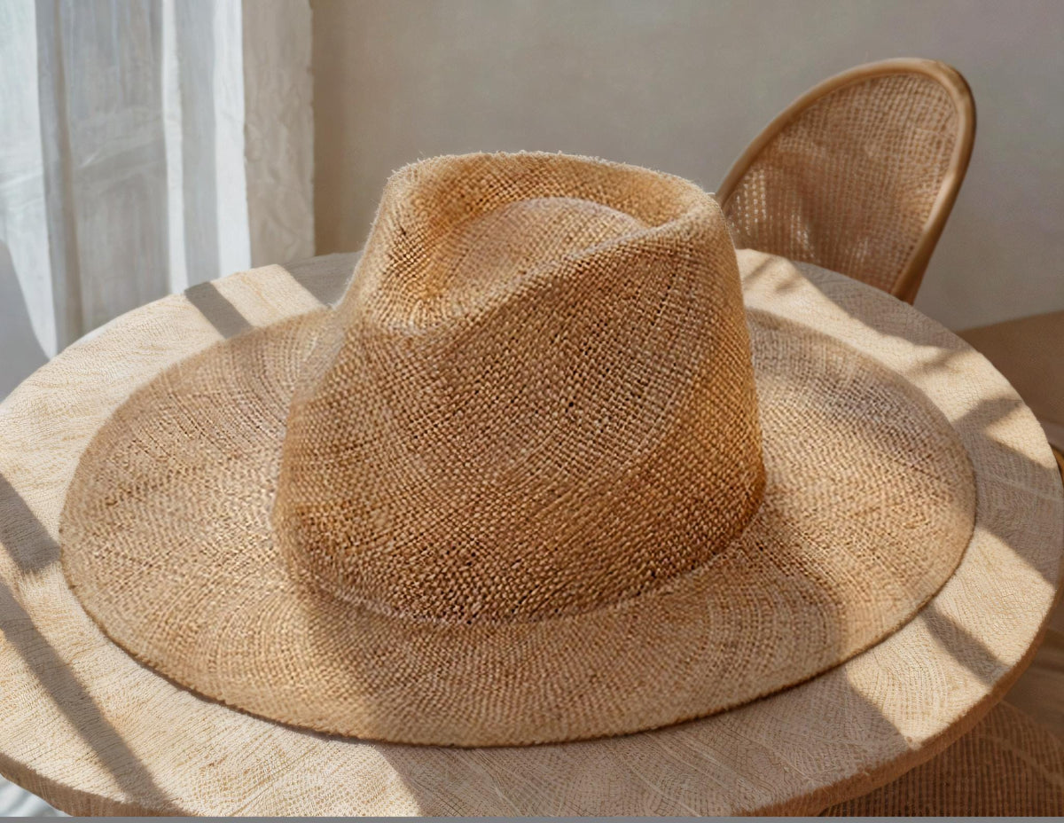 Wide brim straw sun hat with natural woven texture, placed on a wooden table in sunlight near a chair. Lightweight, stylish summer hat for beach, outdoor UV protection, vacation, honeymoon, garden, or as a thoughtful gift for her mom.