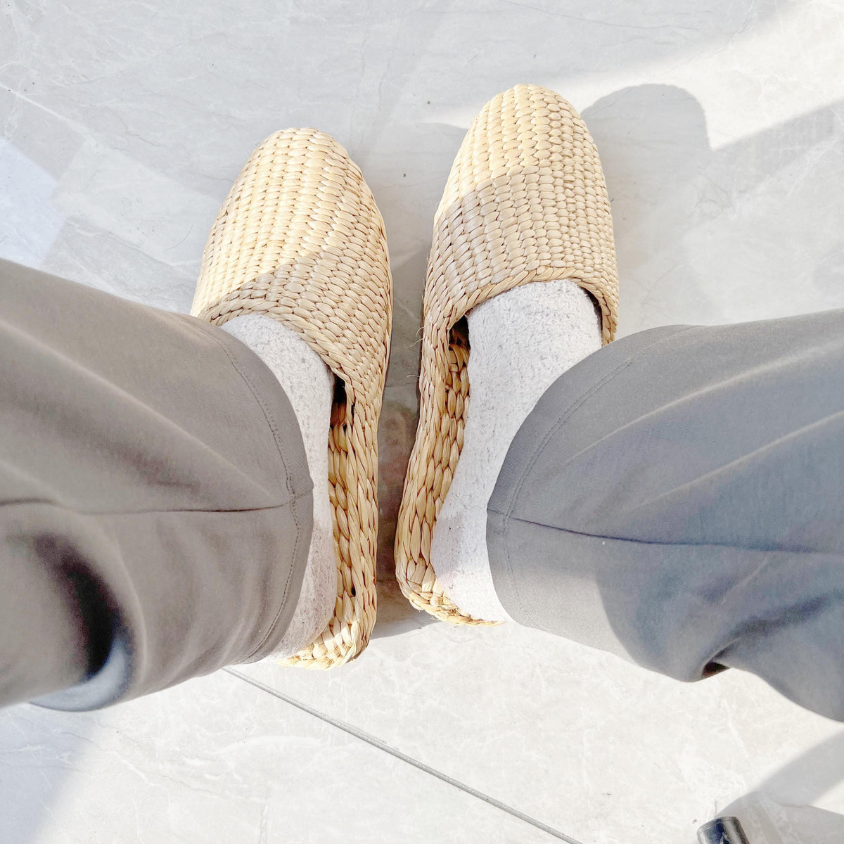 Person wearing handmade straw slippers with closed toe and woven texture, paired with light socks and gray pants. Natural beige color, lightweight and breathable, perfect for house shoes.