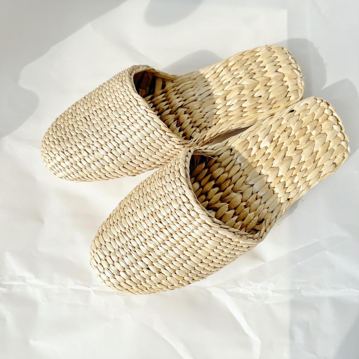 Pair of handmade straw slippers with closed toe and woven texture, displayed on a white background. Natural beige color, lightweight and breathable, ideal for house shoes.