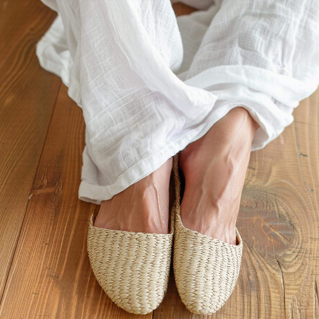 Woman wearing handmade straw slippers with closed pointed toe and woven texture, paired with white linen pants. Natural beige color, lightweight and breathable, ideal for house shoes or summer sandals.