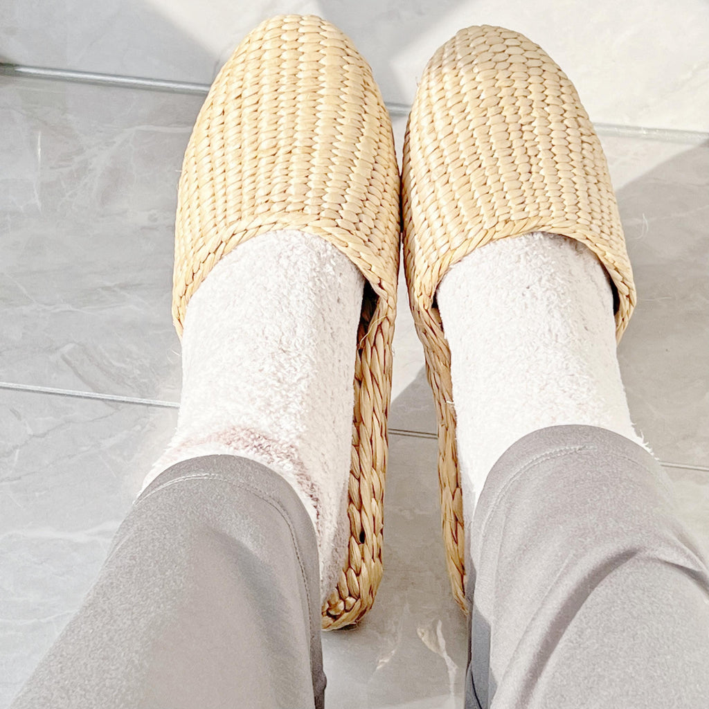 Person wearing handmade straw slippers with closed toe and woven texture, paired with light-colored socks and gray pants. Natural beige color, lightweight and breathable, ideal for house shoes.