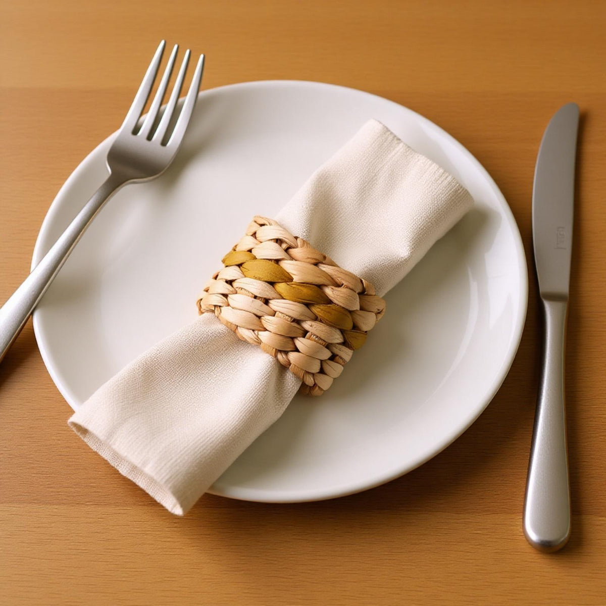 A white plate with a neatly folded napkin, silverware, and a woven napkin ring on a wooden table.