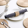 A neatly arranged table setting with a white plate, silverware, and a rolled-up napkin on a woven placemat.