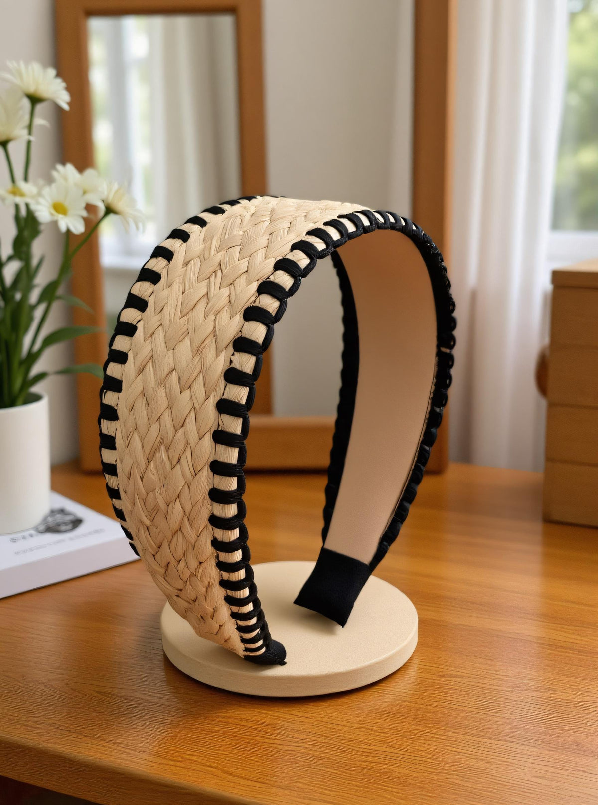 A beige woven headband with black stripes is displayed on a wooden table, accompanied by a vase of white flowers and a book.