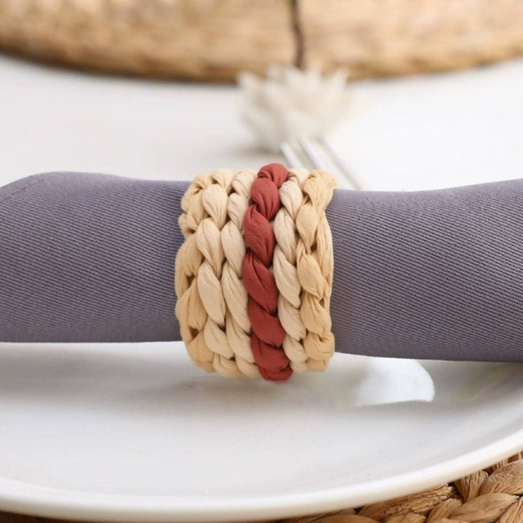 A crocheted napkin ring with a mix of red and beige yarn is placed on a white plate, with a blurred background suggesting a dining setting.