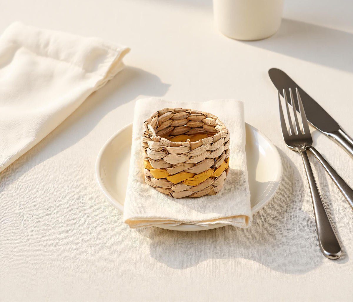 A woven basket with a yellow and brown pattern sits on a white plate, accompanied by a white napkin, silverware, and a white mug on a white tablecloth.