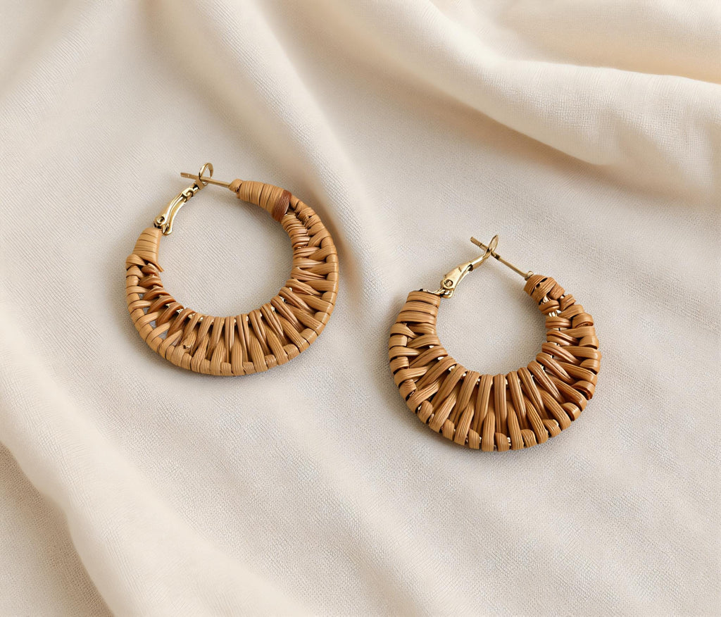 Two beige woven earrings are displayed on a white fabric background.