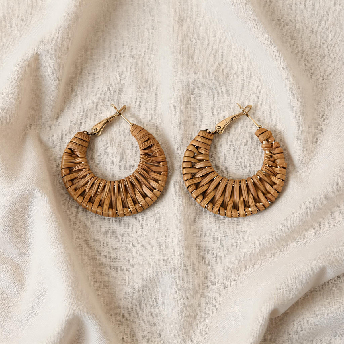 A pair of brown woven hoop earrings is displayed on a white fabric background.