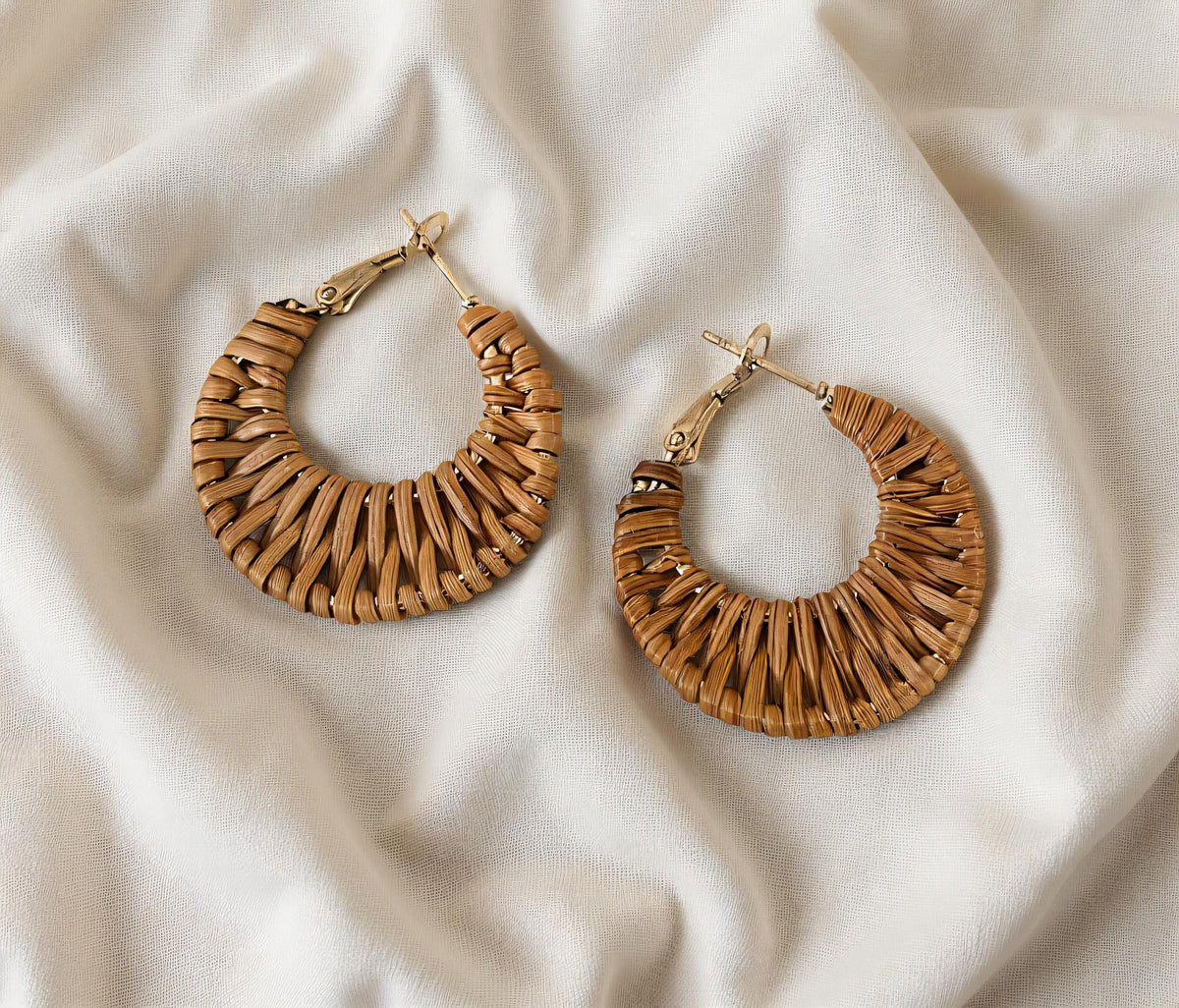 Two gold earrings with woven designs are placed on a white fabric background.
