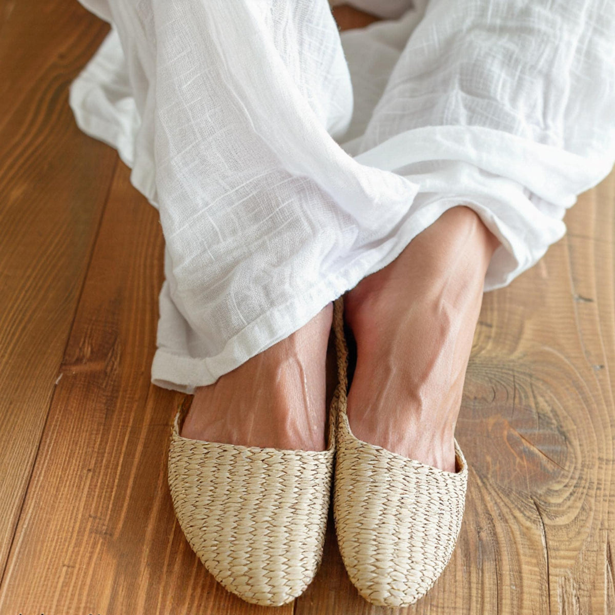 Woman wearing handmade straw slippers with closed pointed toe and woven texture, paired with white linen pants. Natural beige color, lightweight and breathable, ideal for house shoes or summer sandals.