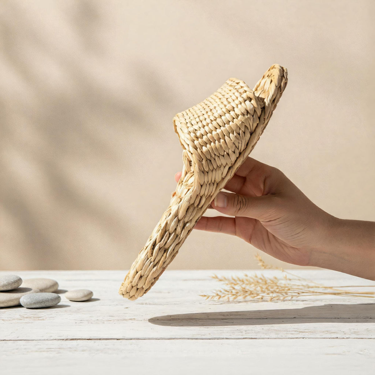 A hand holding a woven straw sandal against a light beige background.