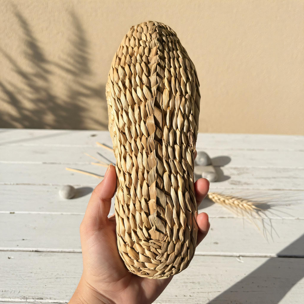A hand holding a woven object resembling a shoe or sandal, with a wooden surface and some wheat stalks in the background.