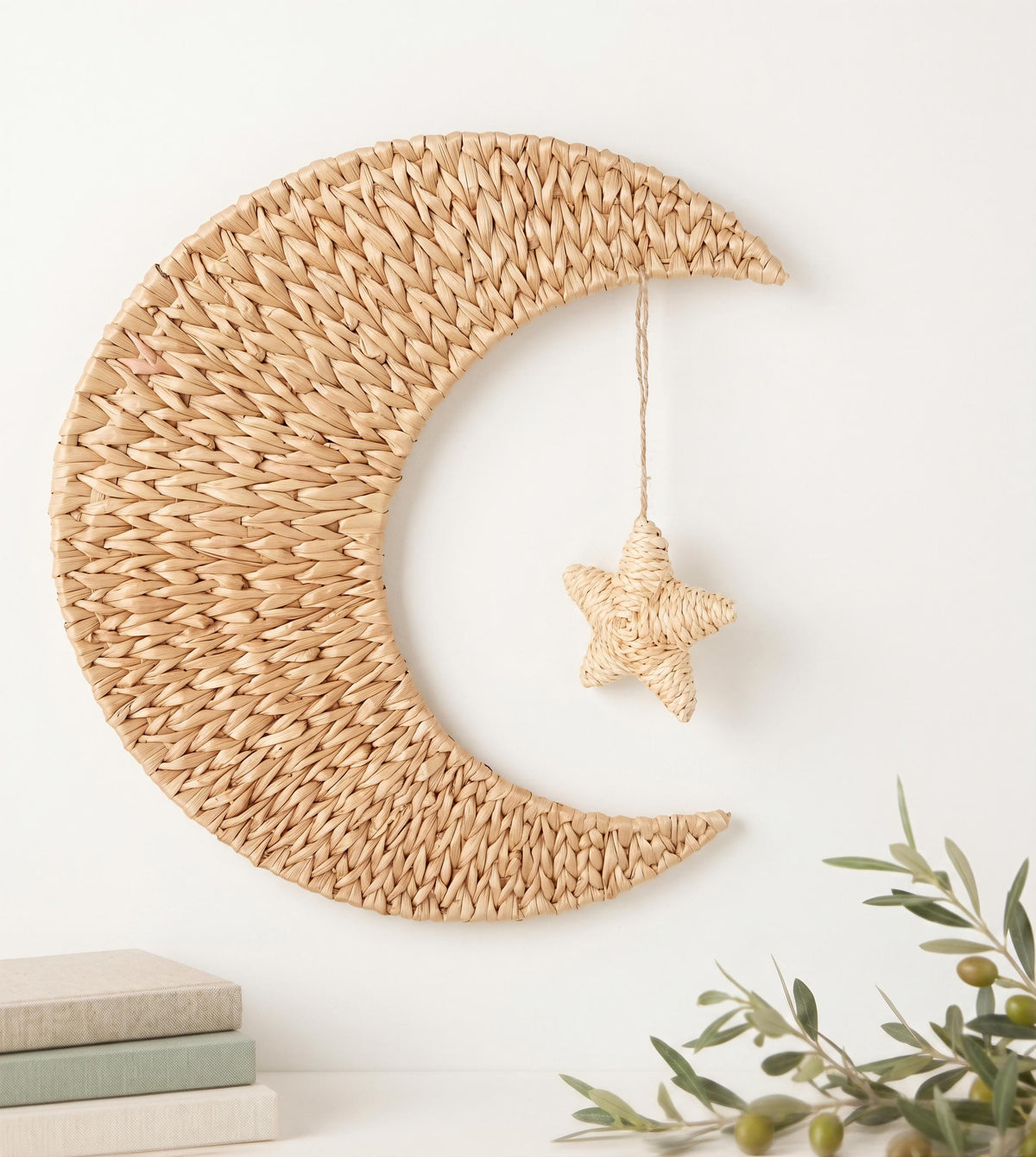 a woven crescent moon-shaped decoration hanging on a white wall, with a small star-shaped ornament hanging from it. The wall is also adorned with a few books and some greenery, creating a cozy and inviting atmosphere.