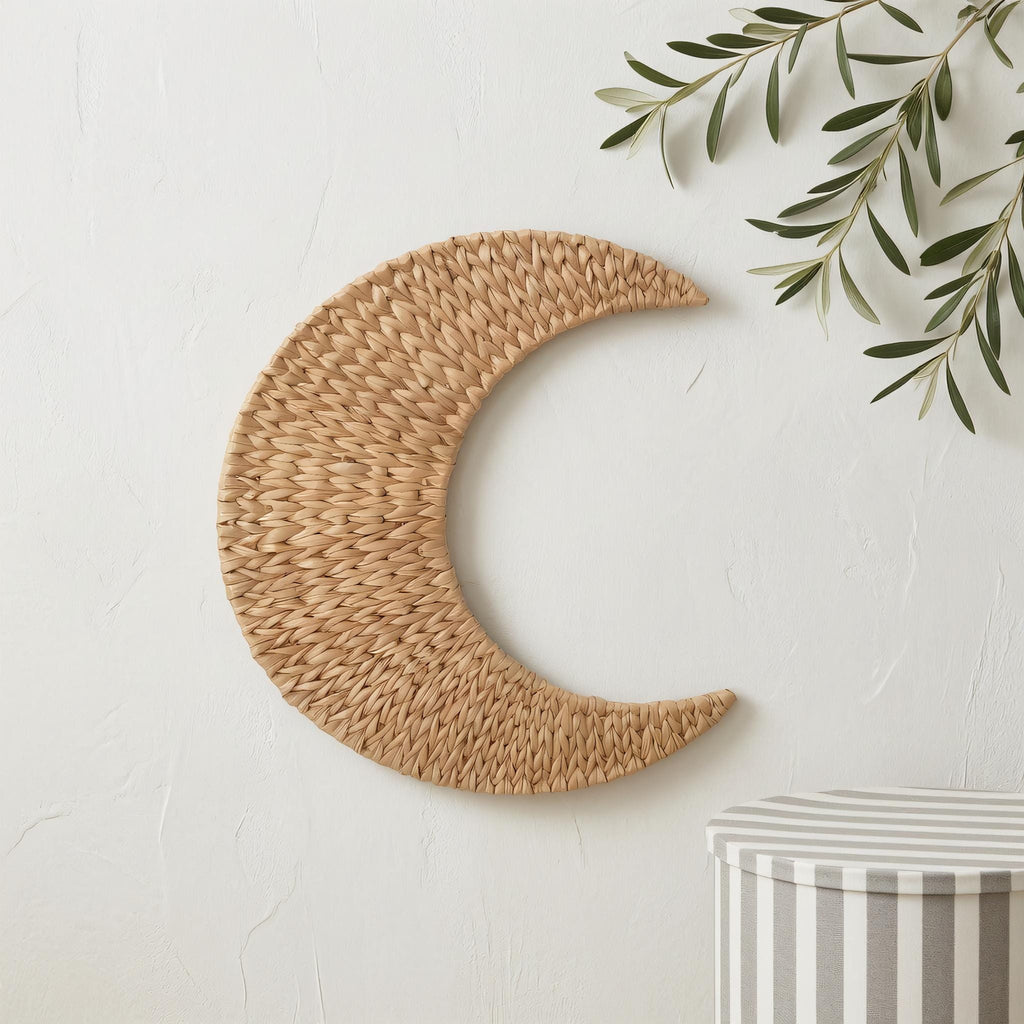 a woven crescent moon-shaped decoration hanging on a white wall, accompanied by a small plant or branch with green leaves.