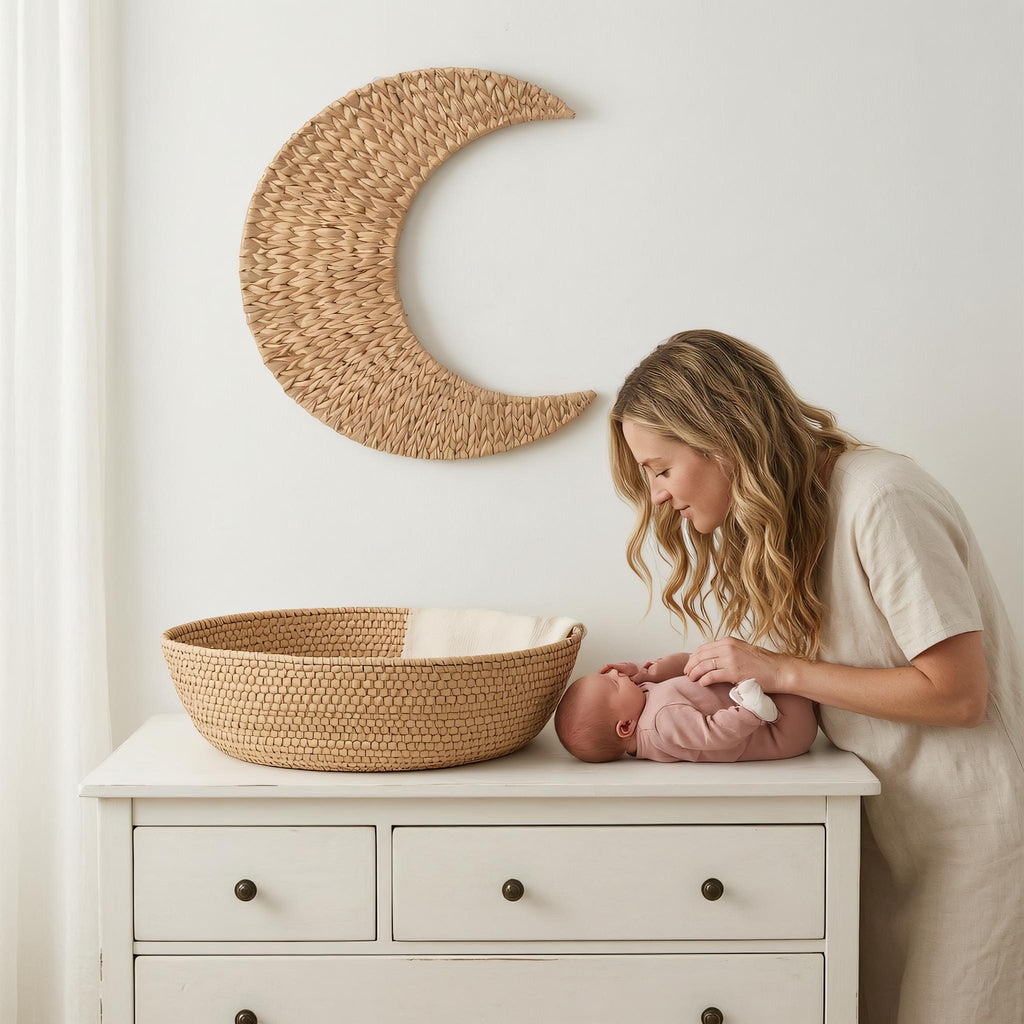 A woman tenderly interacting with a baby in a white dress, standing next to a wooden dresser with a woven basket.