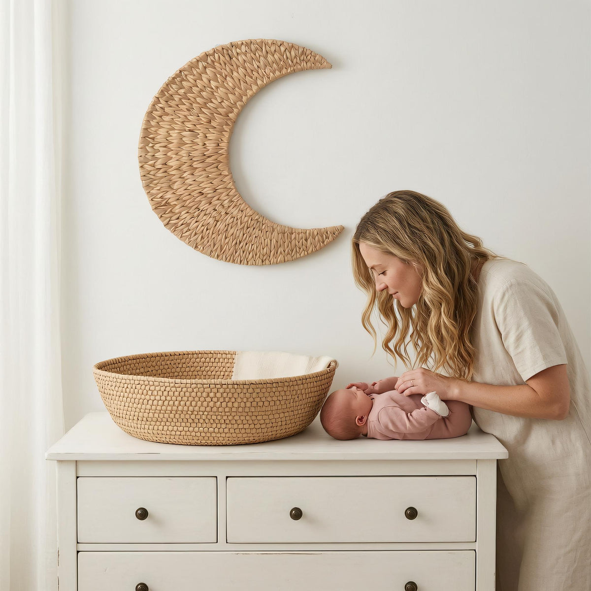 A woman tenderly interacting with a baby in a white dress, standing next to a wooden dresser with a woven basket.