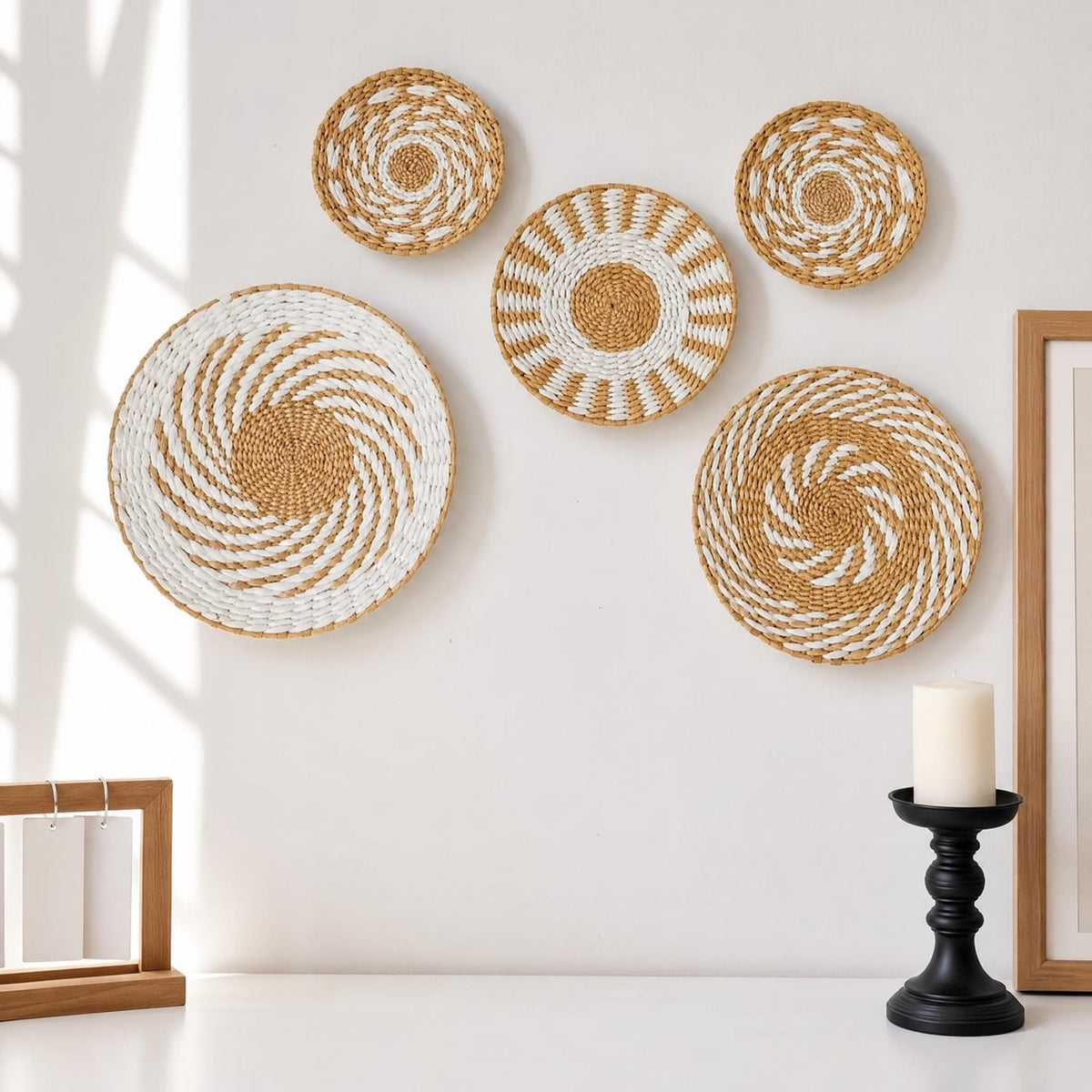 a white wall with five woven baskets of varying sizes and shapes, including a large circular basket and a smaller circular basket. A black candle holder is placed on a white surface, and a wooden frame is visible on the left side of the image.