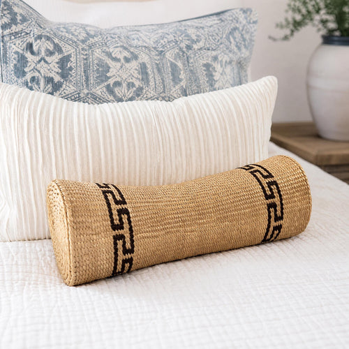 A woven pillow with a black geometric pattern rests on a white bedspread, accompanied by a blue and white patterned pillow and a white vase on a wooden nightstand.