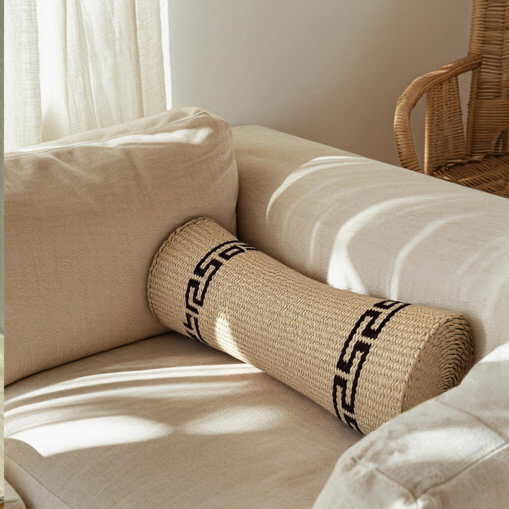 A beige sofa with a woven pillow featuring a black Greek key pattern, placed in a room with a wicker chair visible in the background.