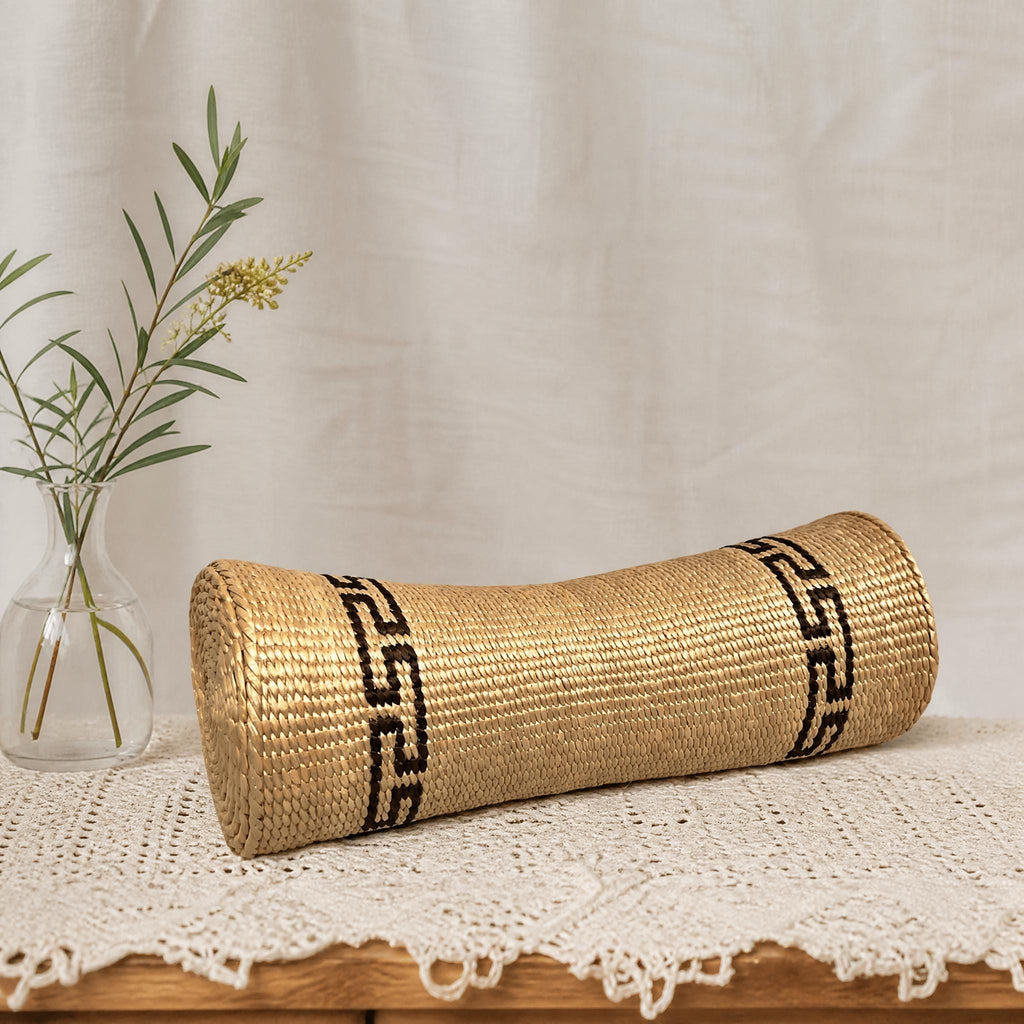 A woven, golden-colored pillow with black geometric patterns rests on a white lace doily, accompanied by a small vase with a plant.