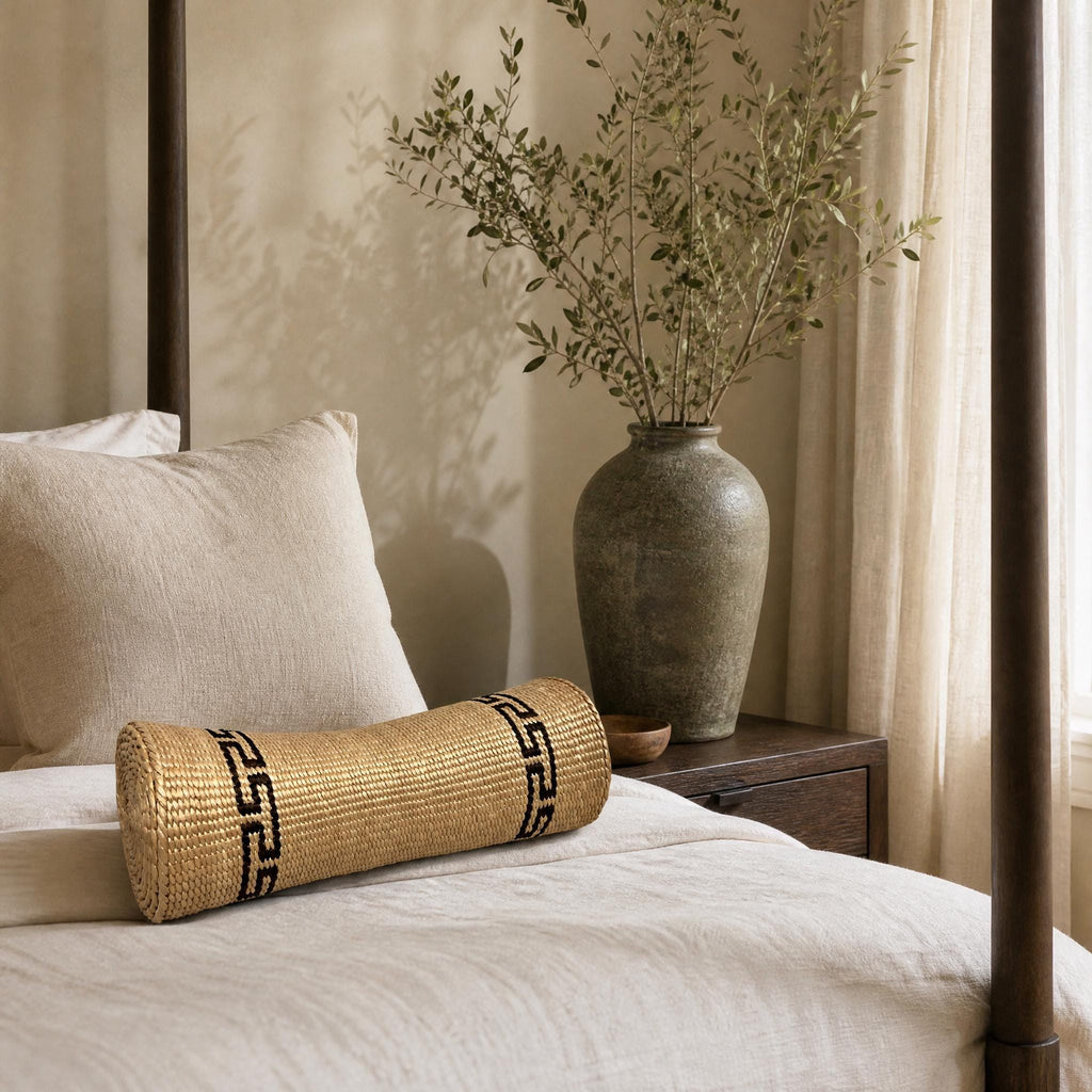 a bedroom with a bed, a wooden nightstand, and a decorative vase with greenery. The bed has a white pillow and a woven pillow on it. The nightstand has a wooden frame and a small vase with greenery on it. The overall color scheme of the room is neutral, with a beige color palette.