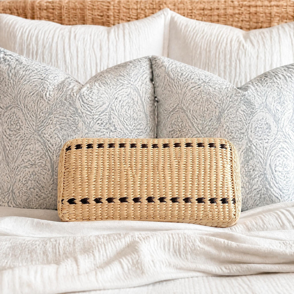 A woven straw bag with black accents rests on a bed with white bedding and patterned pillows.